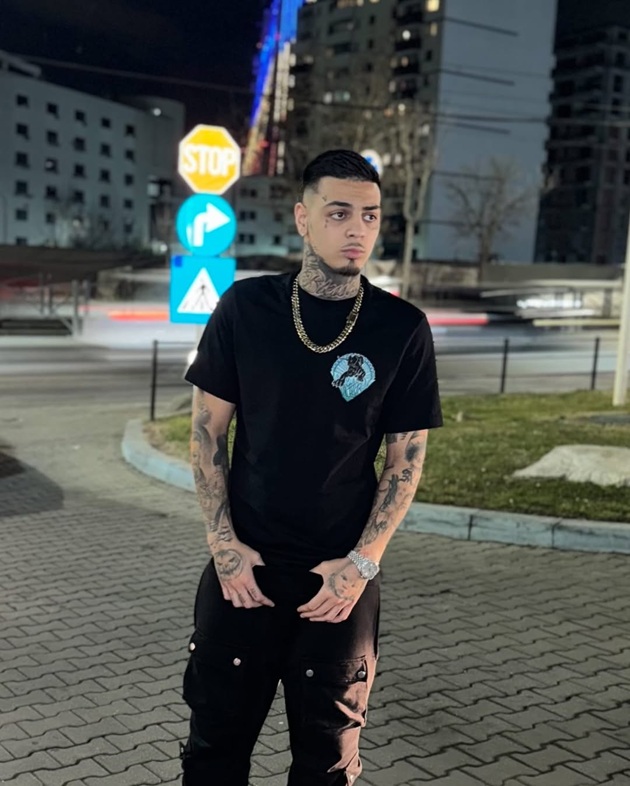 Albert NBN ținându-se cu mâinile de pantaloni și uitându-se într-o parte, îmbrăcat în negru și purtând un lanț masiv la gât