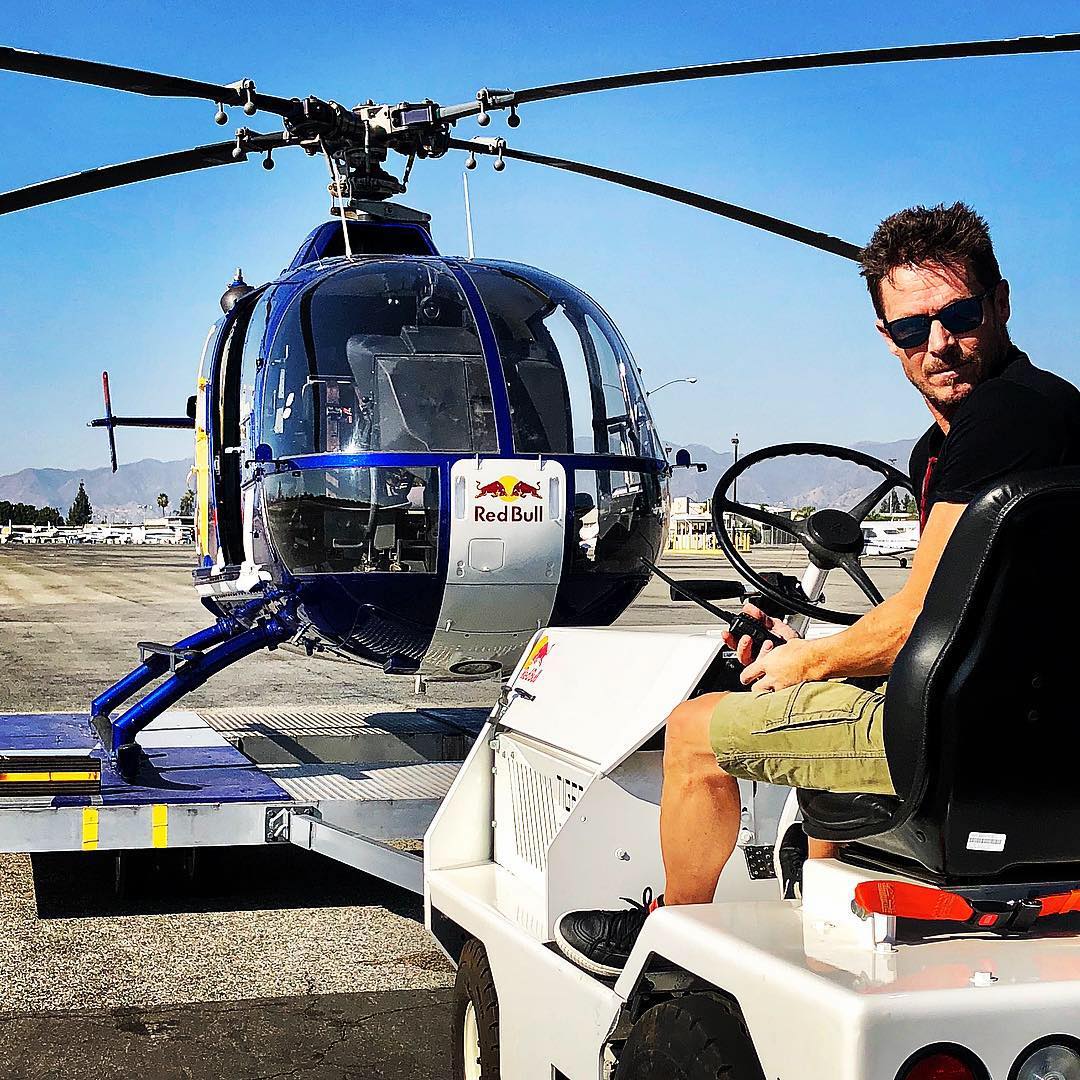 Felix Baumgartner cu ochelarii de soare la ochi, stând într-un autovehicul pe pistă, lângă un elicopter