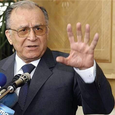 Ion Iliescu în timpul unui interviu, îmbrăcat în costum negru și cămașă albă, purtând o cravată cu buline și făcând un semn cu mâna