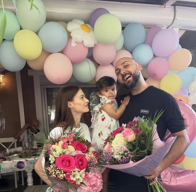 Teodora și Alex Marcu ținând în mână buchete de flori, înconjurați de baloane, stând alături de fiica lor pe 31 iulie 2024, când micuța Emma a împlinit 2 ani