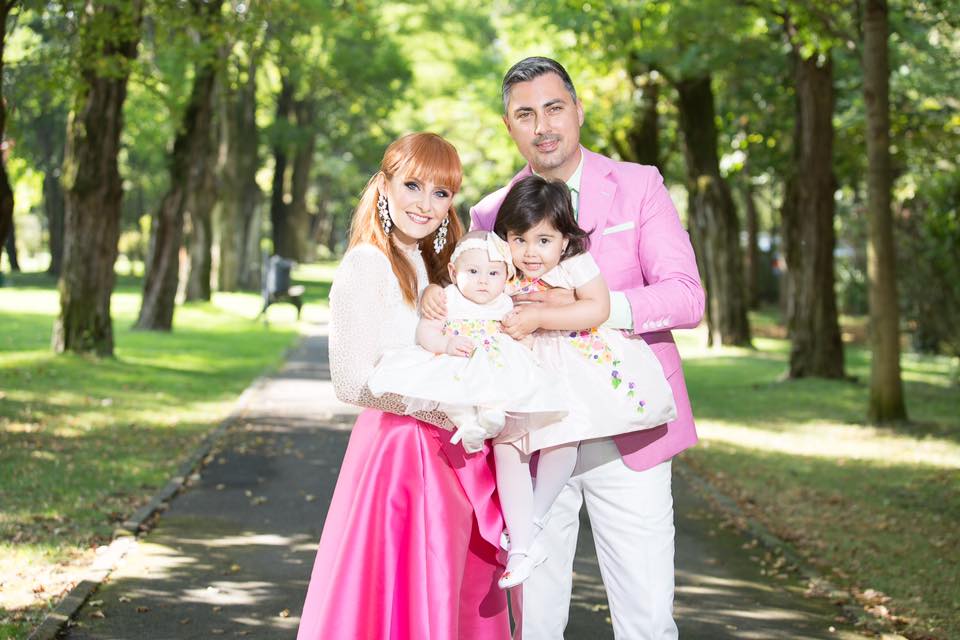 Alina Sorescu și Alexandru Ciucu, în urmă cu mai mulți ani, ținându-le în brațe pe fiicele lor, Carolina și Raisa, pe aleea unui parc, în timp ce erau îmbrăcați în ținute elegante, pregătiți de botezul mezinei