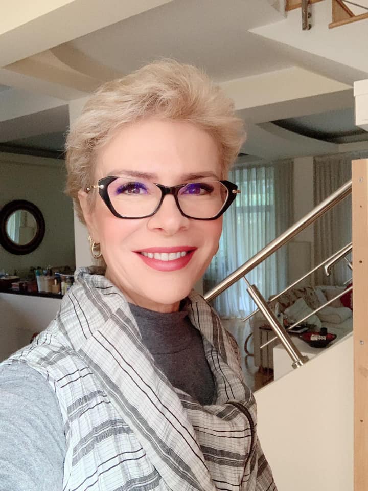 Teo Trandafir zâmbitoare, cu ochelarii la ochi, făcându-și un selfie lângă o scară, îmbrăcată în mai multe nuanțe de gri