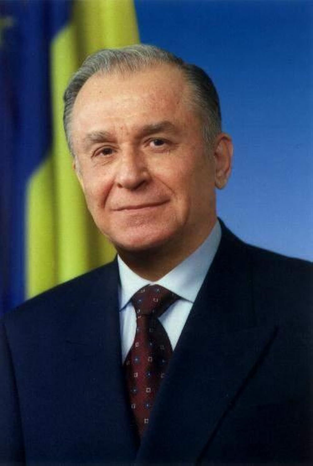 Ion Iliescu, având steagul României în spate, îmbrăcat în costum negru, cămașă albă și cravată maro cu pătrățele albe