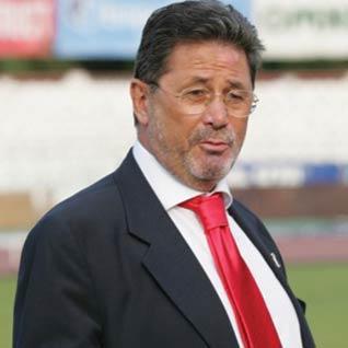 Cornel Dinu pe terenul de fotbal, îmbrăcat în costum închis la culoare, purtând o cămașă albă și o cravată roșie