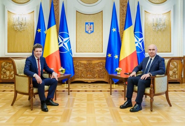 Nicușor Dan și Ilie Bolojan, stând pe două scaune alăturate, în timpul unei întâlniri oficiale, cu steagurile României, Uniunii Europene și NATO în spate