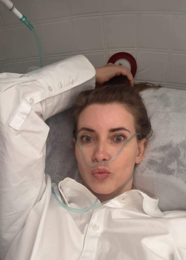 Iulia Albu în cămașă albă, cu părul prins, stând pe patul de spital, conectată la oxigen
