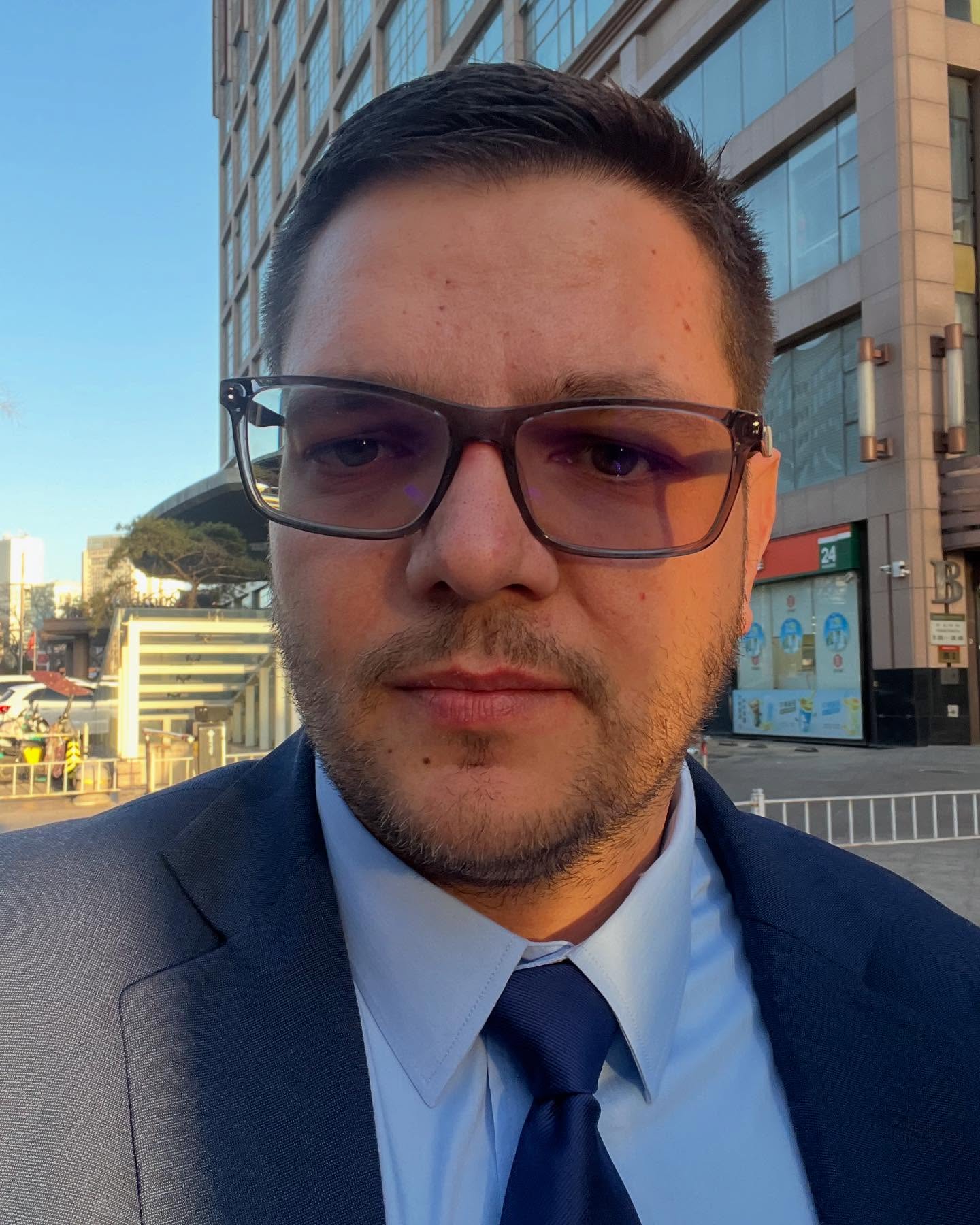 Victor Dăncilă, fiul adoptat de Viorica Dăncilă și de soțul ei, Cristinel Dăncilă, în costum închis la culoare, cu cămașă albă, cravată albastră și ochelari de vedere la ochi
