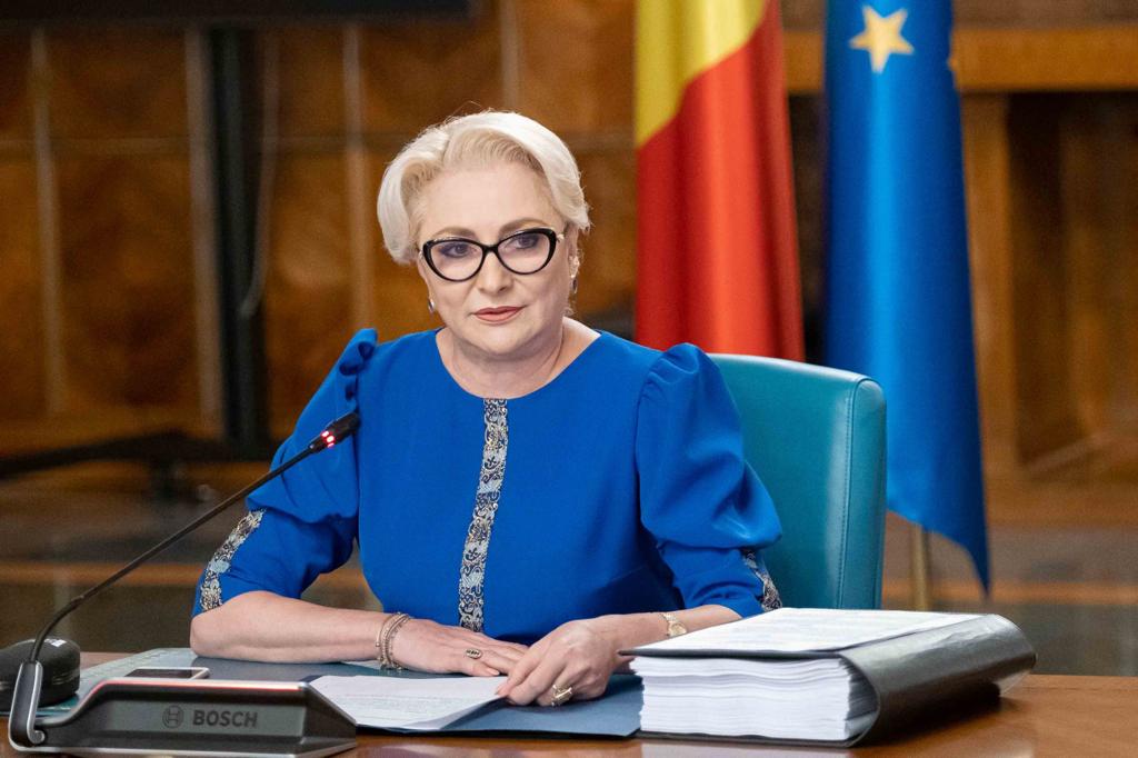 Viorica Dăncilă într-o rochie albastră cu detalii aurii, stând la birou, lângă un microfon, vorbind în fața unui public