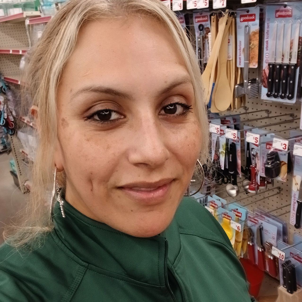 Consuelo Bernabe făcându-și un selfie printre rafturile unui magazin, zâmbitoare, îmbrăcată într-o bluză de trening verde, purtând cercei mari în urechi