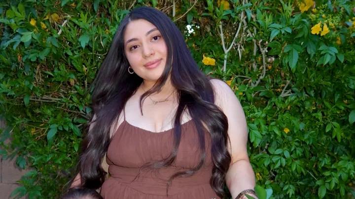 Yarely Ashley Hermosillo zâmbitoare, într-o rochie maro cu bretele subțiri, purtând bijuterii aurii
