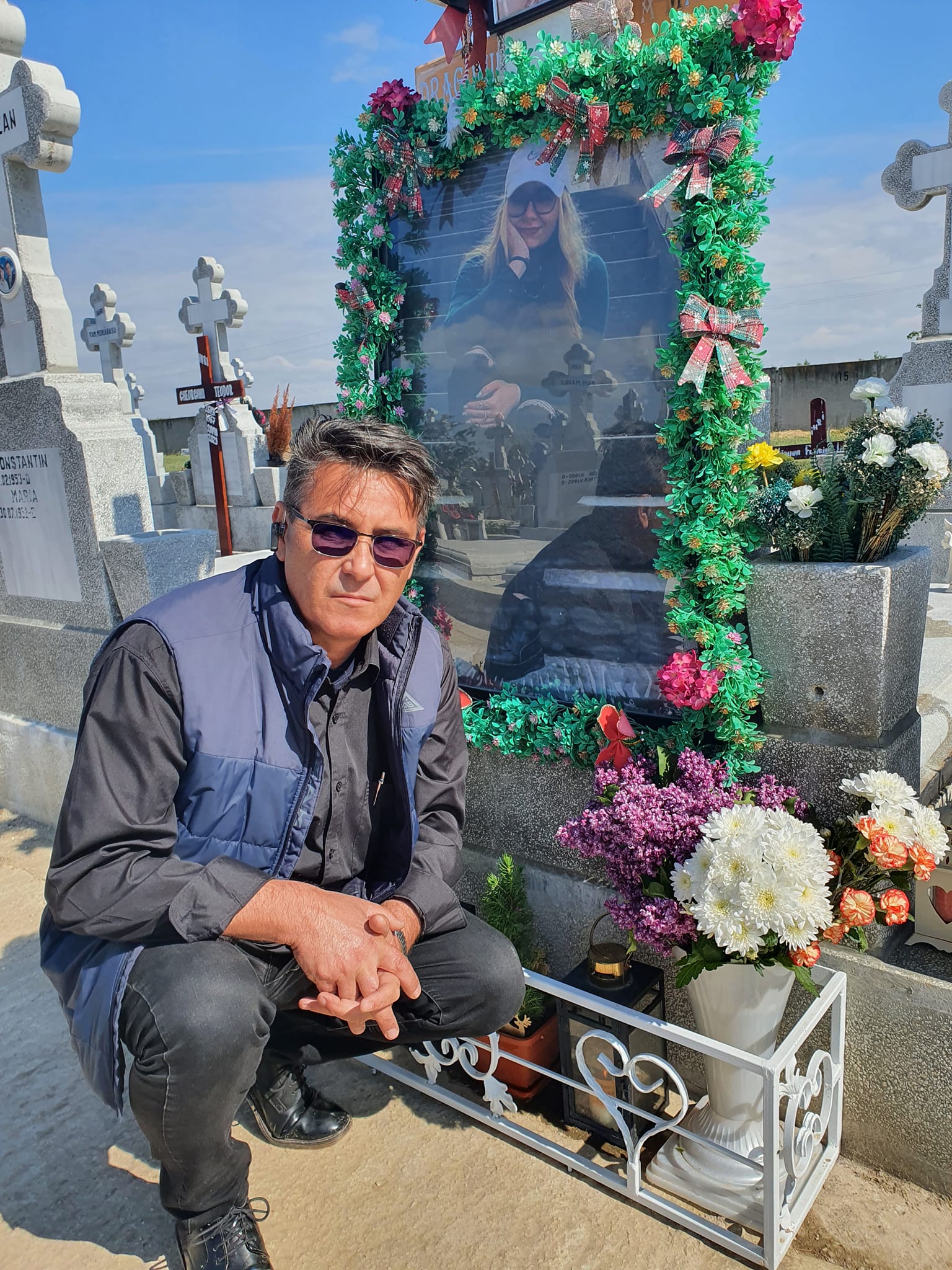 Cătălin Dragomir stând pe vine lângă mormântul fiicei sale, care este acoperit de flori,
