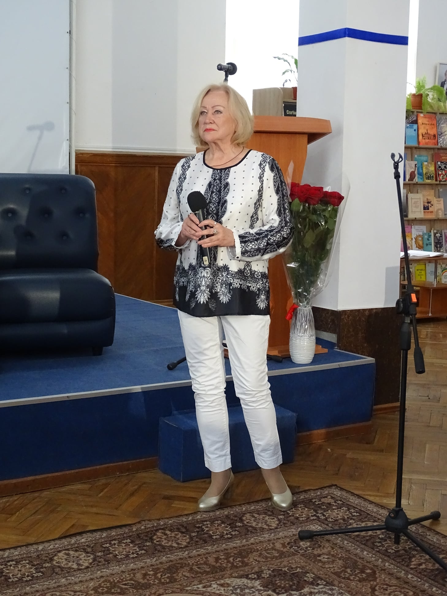 Margareta Ivănuș îmbrăcată în alb și negru, cu microfonul în mână, stând în fața unei scene