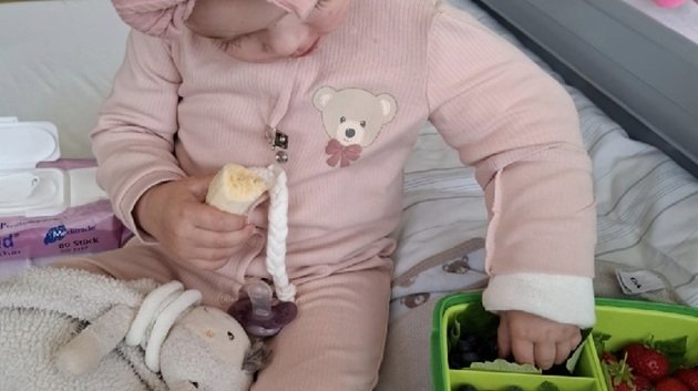 Micuța Hannah pe patul de spital, îmbrăcată într-o salopetă roz, mâncând fructe, după ce s-a trezit din comă