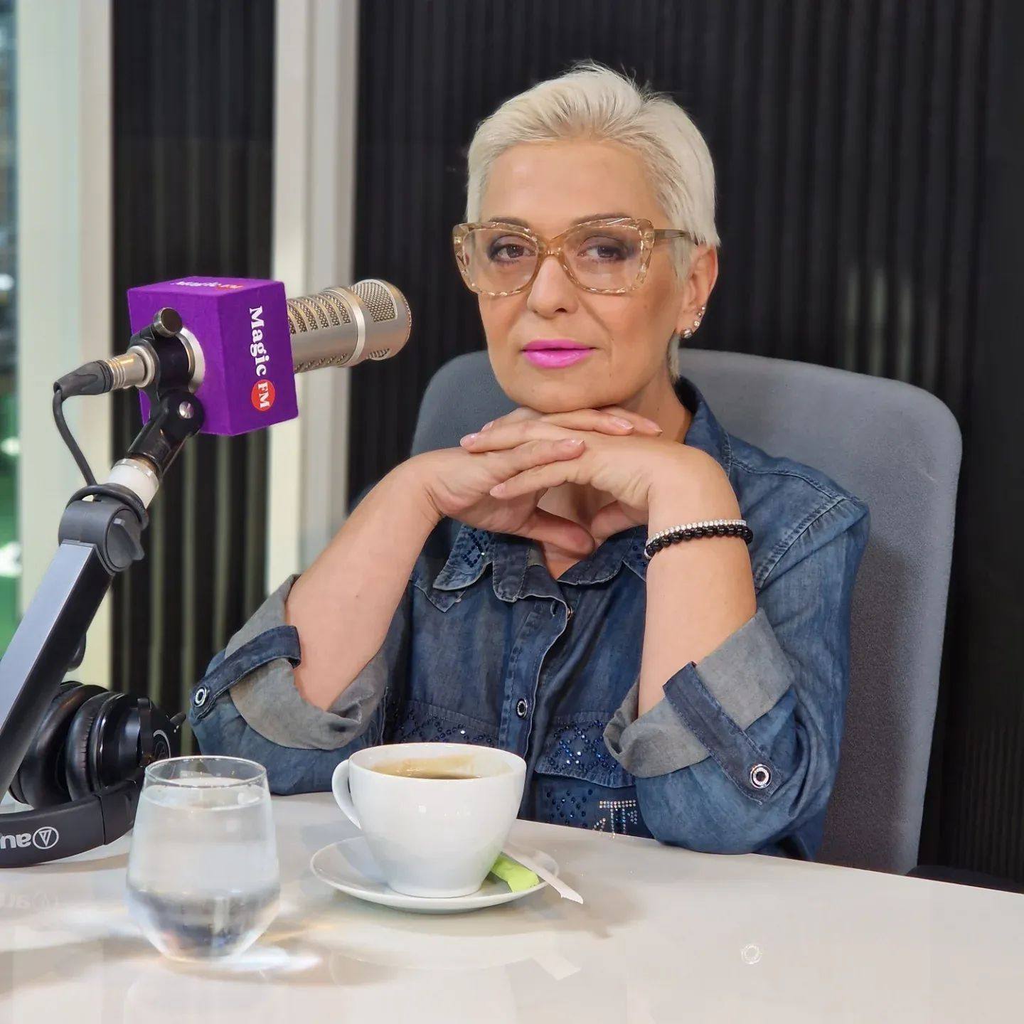 Monica Anghel, la microfonul, în studioul Magic FM, cu o cafea și o apă în față, purtând ochelari de vedere