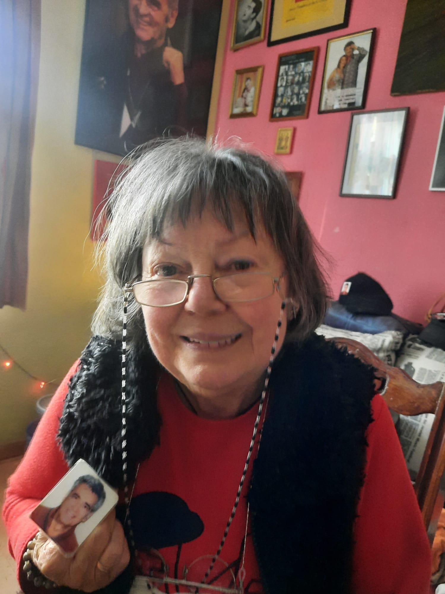 Anca Pandrea ținând în mână o fotografie cu Iurie Darie, în timp ce stă în camera care are pereții împodobiți cu tablouri cu el