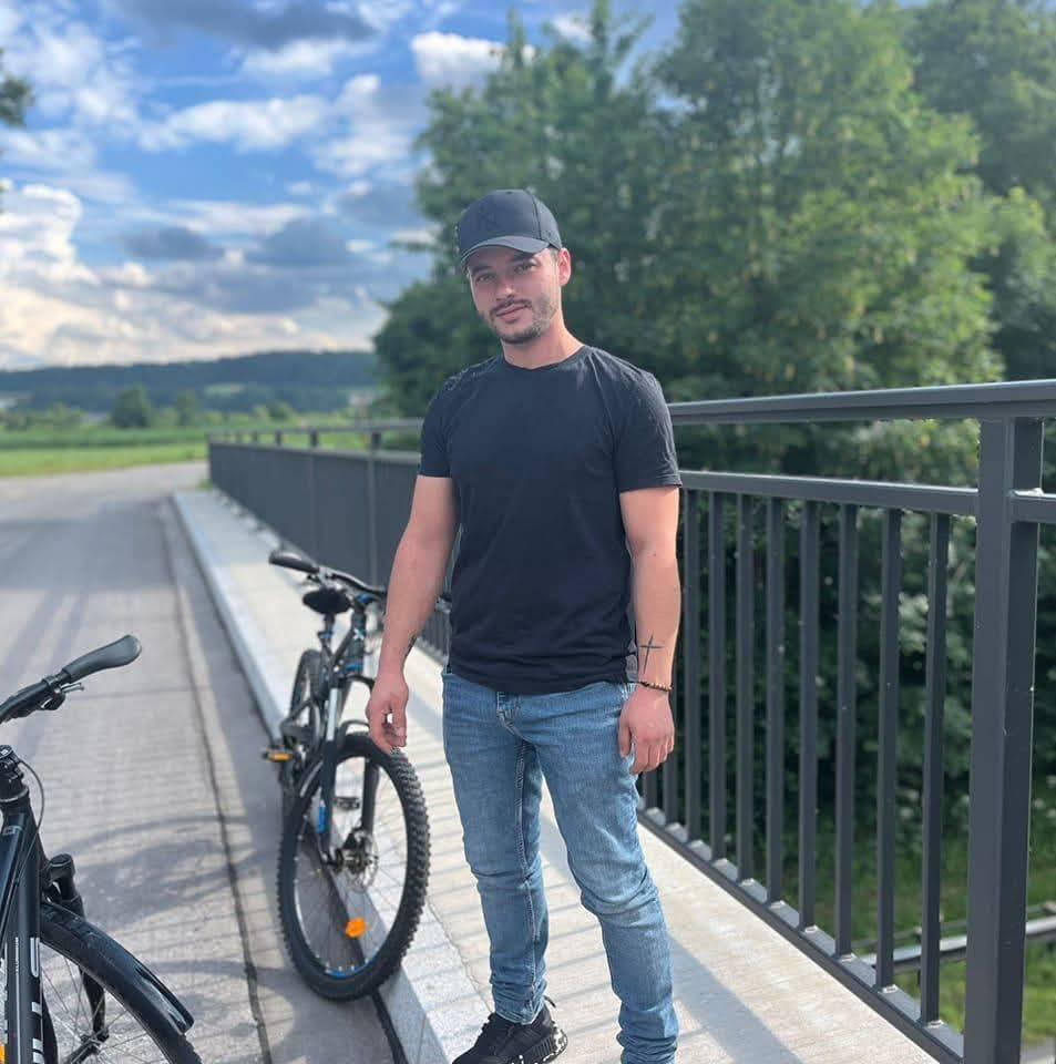Radu Adrian Florin la plimbare cu bicicleta, stând pe marginea drumului, pe un pod, într-o zonă de munte, îmbrăcat în blugi, tricou negru și purtând pe cap o șapcă neagră