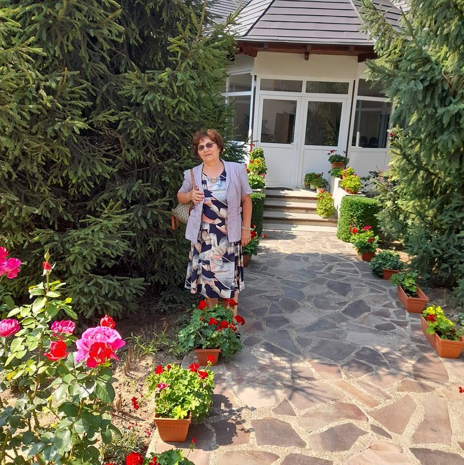 Liliana Aglaia Grădinaru, mama Mirabelei Grădinaru, îmbrăcată elegant, în rochie cu imprimeuri, stând pe o alee plină de flori și copaci verzi