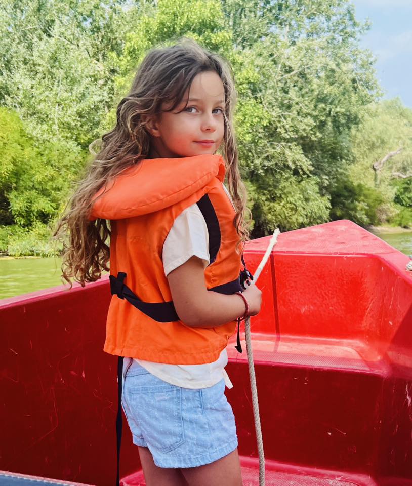 Eva Maria, fiica Elenei Udrea, la o plimbare cu barca în Delta Dunării, îmbrăcată în blugi scurți, tricou alb și cu vesta de salvare pe ea.