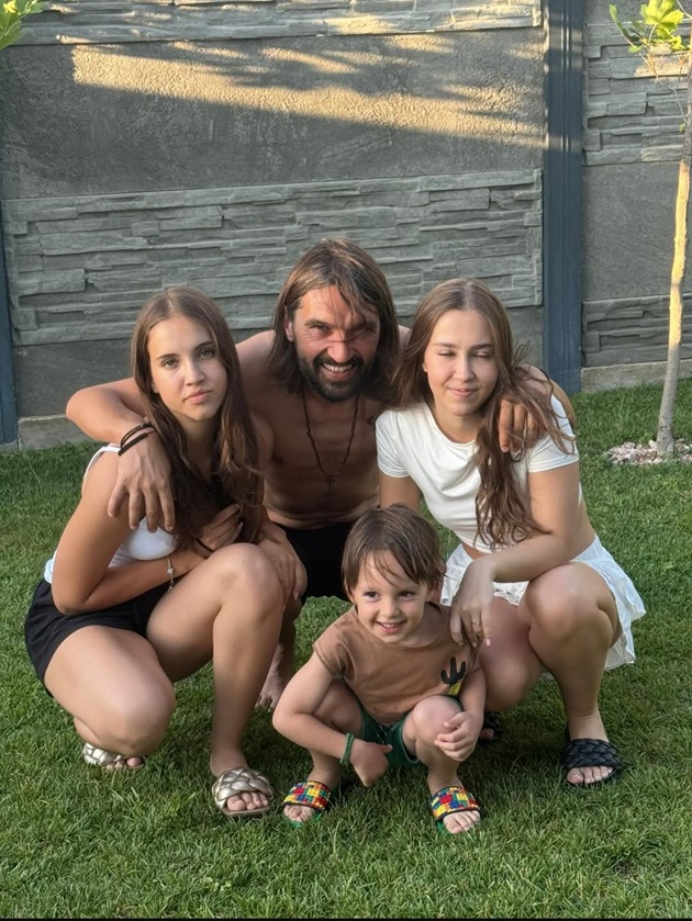 Dan Alexa zâmbitor, stând pe iarbă și îmbrățișându-și cei trei copii