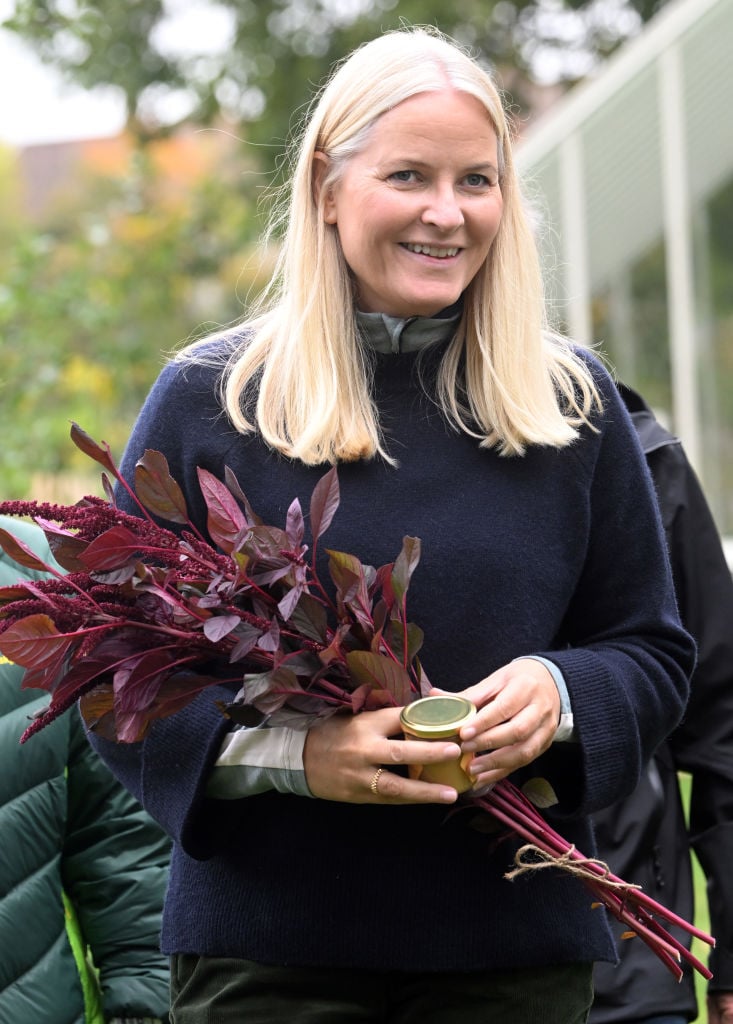 Prințesa Moștenitoare a Norvegiei, Mette‑Marit, zâmbitoare, îmbrăcată în albastru închis, ținând în brațe un buchet de flori și un borcan de miere în mâini