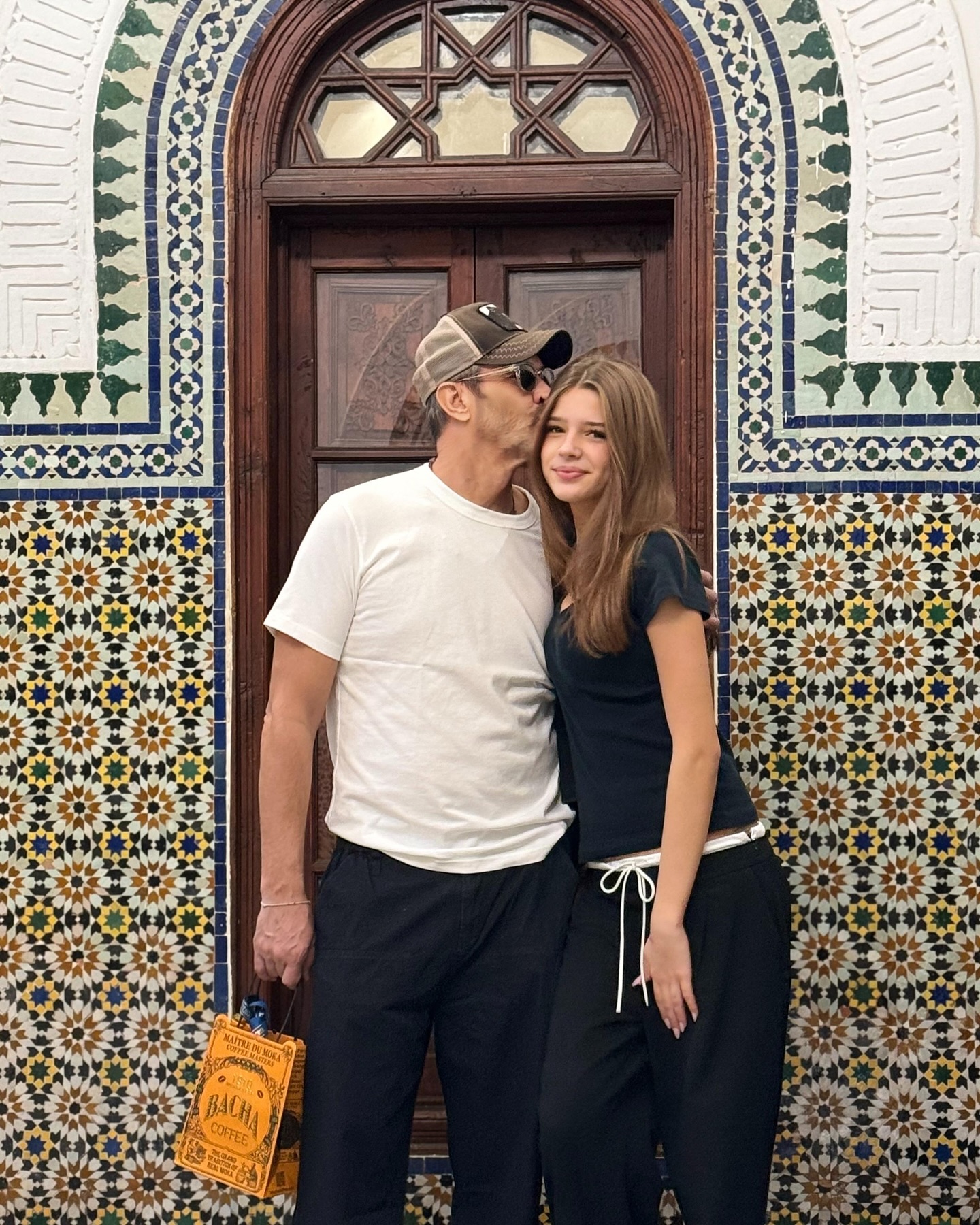 Nicolai Tand sărutând-o pe fiica lui, Ilona Tand, pe frunte, în fața unor pereți cu mozaic și a unei uși maro