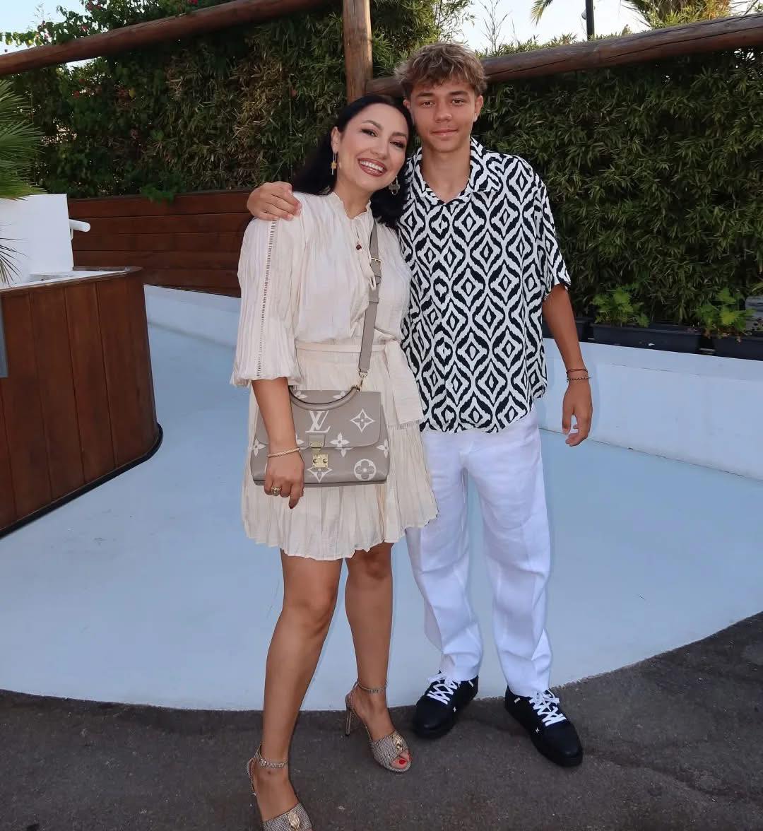 David Măruță zâmbitor, îmbrăcat în alb și negru, ținând-o în brațe pe mama lui, Andra, îmbrăcată într-o rochie de culoarea untului, încălțată cu pantofi cu toc nude și ținând pe umăr o geantă Louis Vuitton