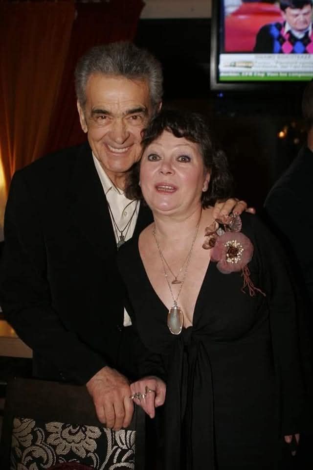 Anca Pandrea zâmbitoare, în brațele lui Iurie Darie, în timp ce erau amândoi îmbrăcați elegant, în negru, la un eveniment.