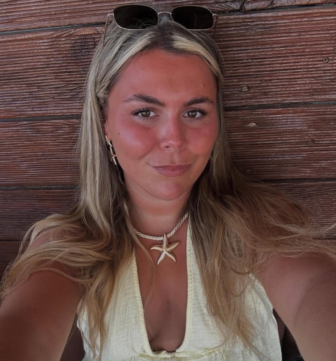 Georgia Taylor făcându-și un selfie lângă un perete, stând cu părul desfăcut și cu ochelarii de soare pe cap, purtând bijuterii cu temarică marină