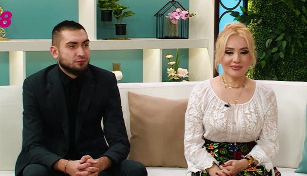 Adriana Ochișanu în costum popular și fiul ei, Cristian Botgros în costum negru, stând împreună pe aceeași canapea, în platoul unei emisiuni