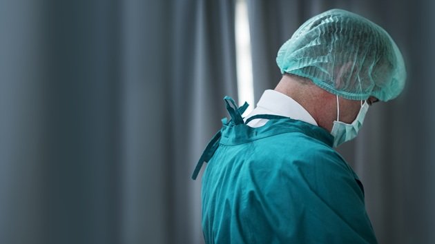 Medic care stă cu spatele la aparatul de fotografiat, îmbrăcat în halat verde, cu bonetă pe cap și mască de protecție pe față