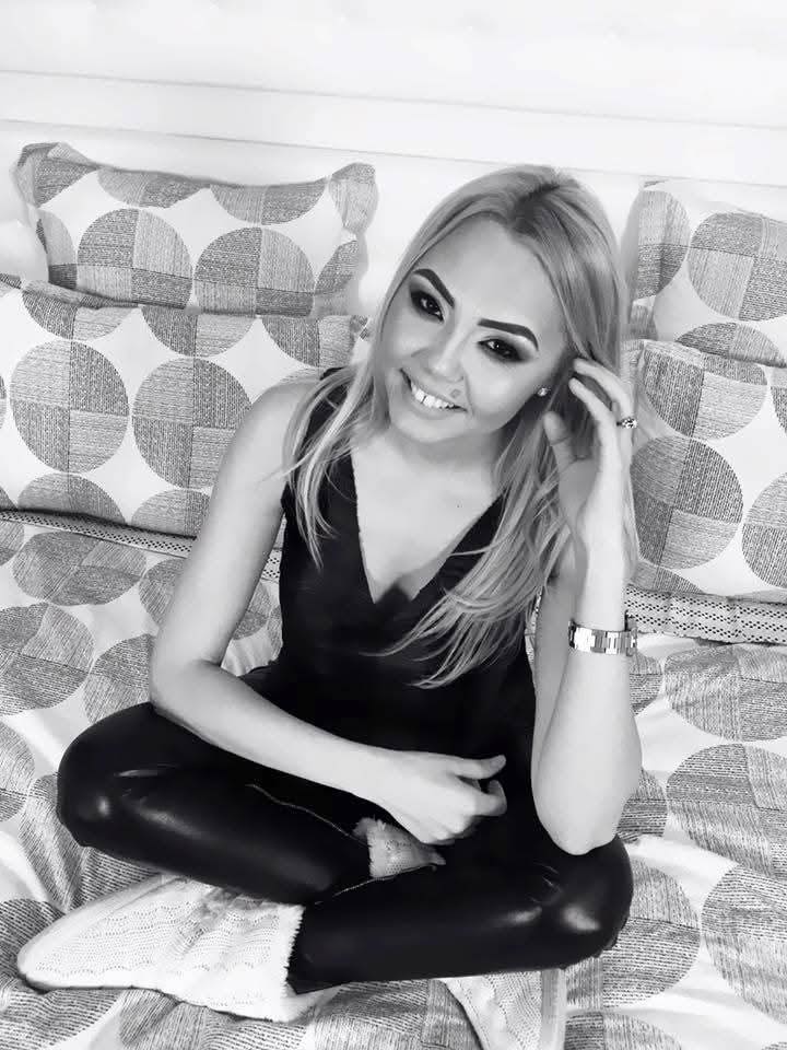 Regretata manelistă Denisa Răducu, zâmbitoare, stând cu picioarele încrucișate pe canapea, îmbrăcată în maoiu negru și pantalini mulați din imitație de piele, cu botoși albi tricotați în picioare