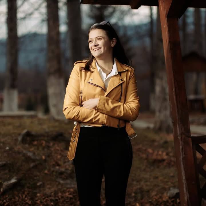 Simona zâmbitoare, îmbrăcată în geacă de piele crem, stând cu mâinile încrucișate într-o pădure