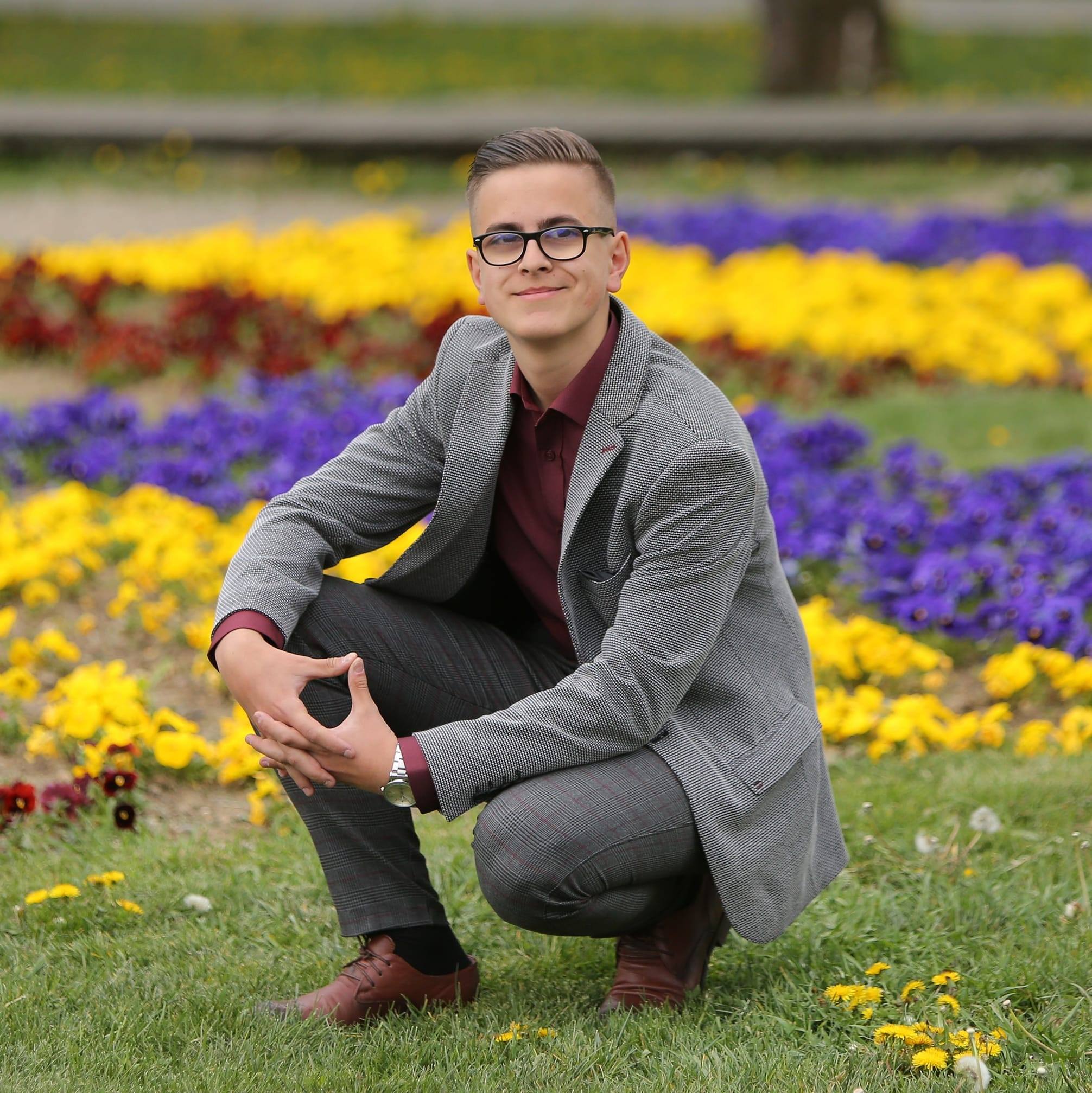 Tudor în costum gri, stând pe vine, lângă multe flori colorate