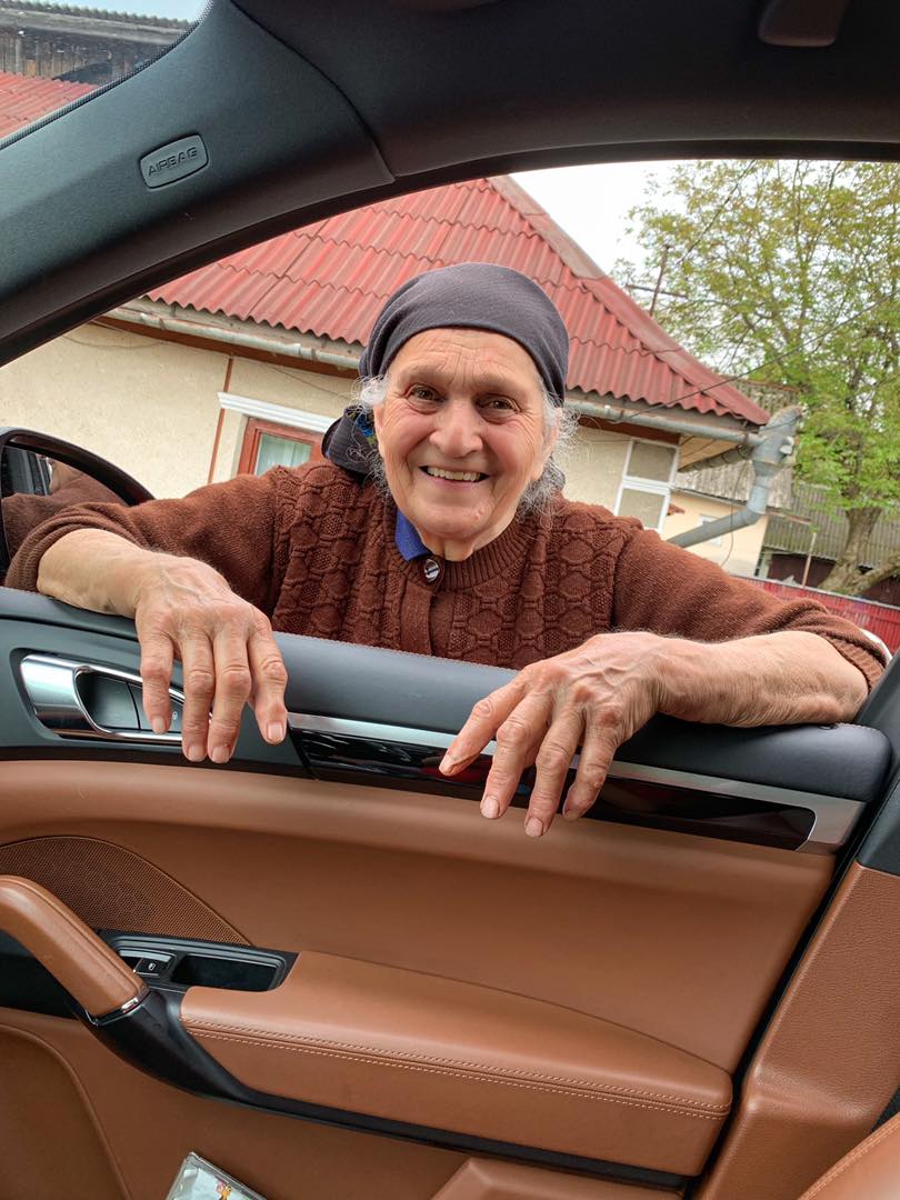 Bunica Vlăduței Lupău zâmbitoare, sprijinindu-se cu mâinile de ușa mașinii pe care o conduce vedeta, bucuroasă că aceasta i-a făcut o vizită