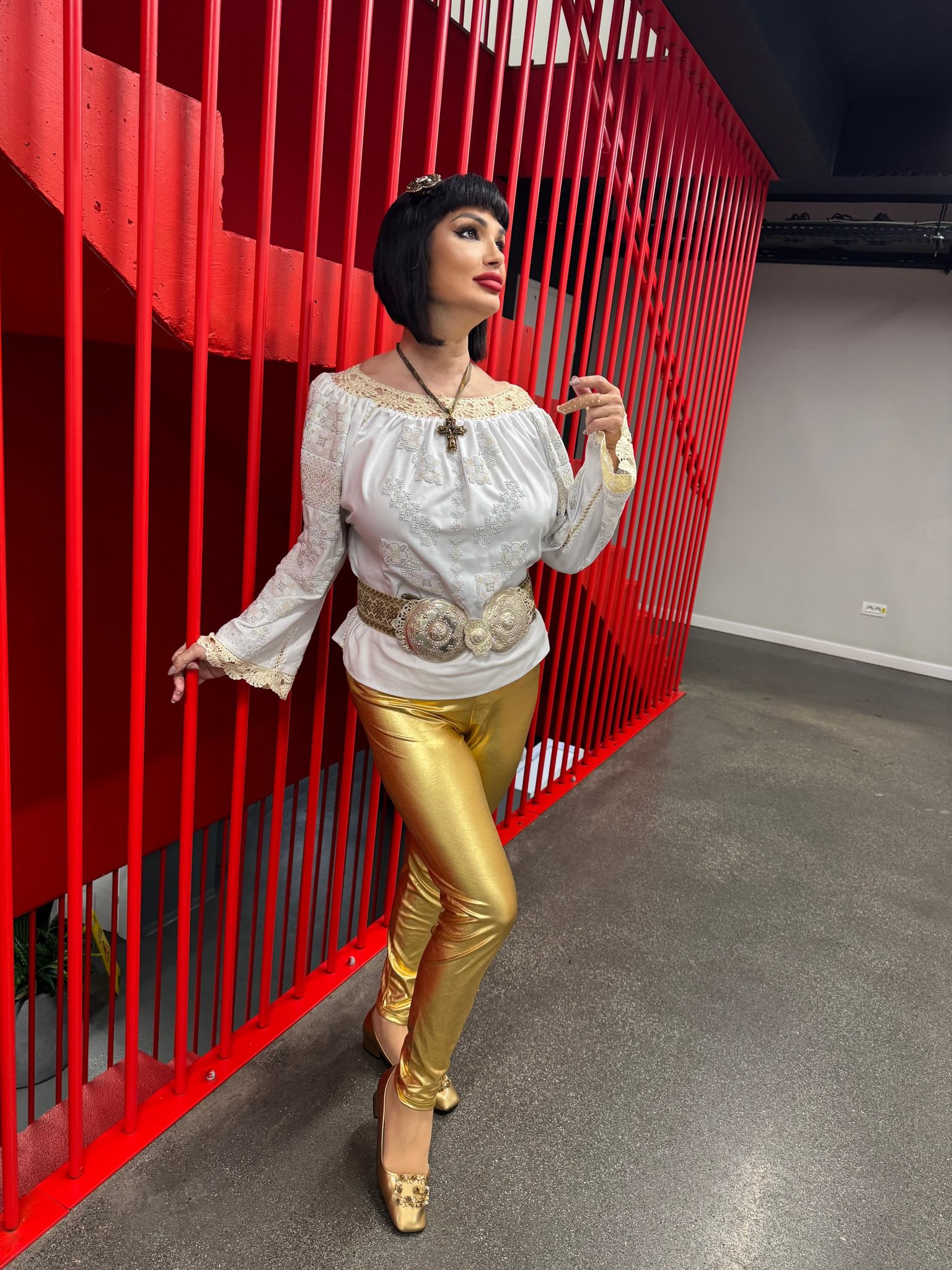 Ozana Barabancea stând lângă o scară cu balustradă roșie, îmbrăcată în pantaloni mulați aurii și bluză albă, mai slabă cu 20 de kilograme
