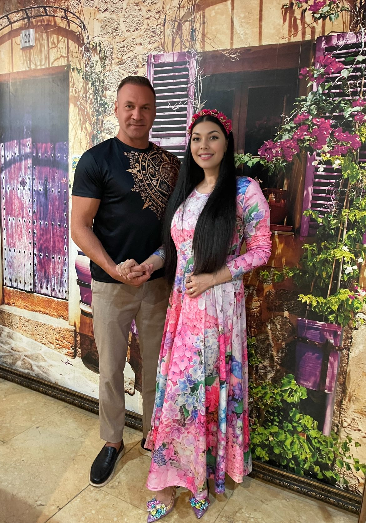 Alin Oprea într-un tricou negru și pantaloni bej, ținând-o de mână pe soția sa Medana, îmbrăcată într-o rochie colorată, cu imprimeu floral, în timp ce stau amândoi lângă un tablou imens, care acoperă un întreg perete