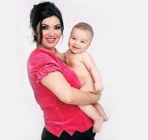 Adriana Bahmuțeanu zâmbitoare, îmbrăcată în pantaloni negri și tricou roz, ținându-l în brațe pe fiul ei cel mare, Maximus Prigoană, care era dezbrăcat în momentul realizării fotografiei