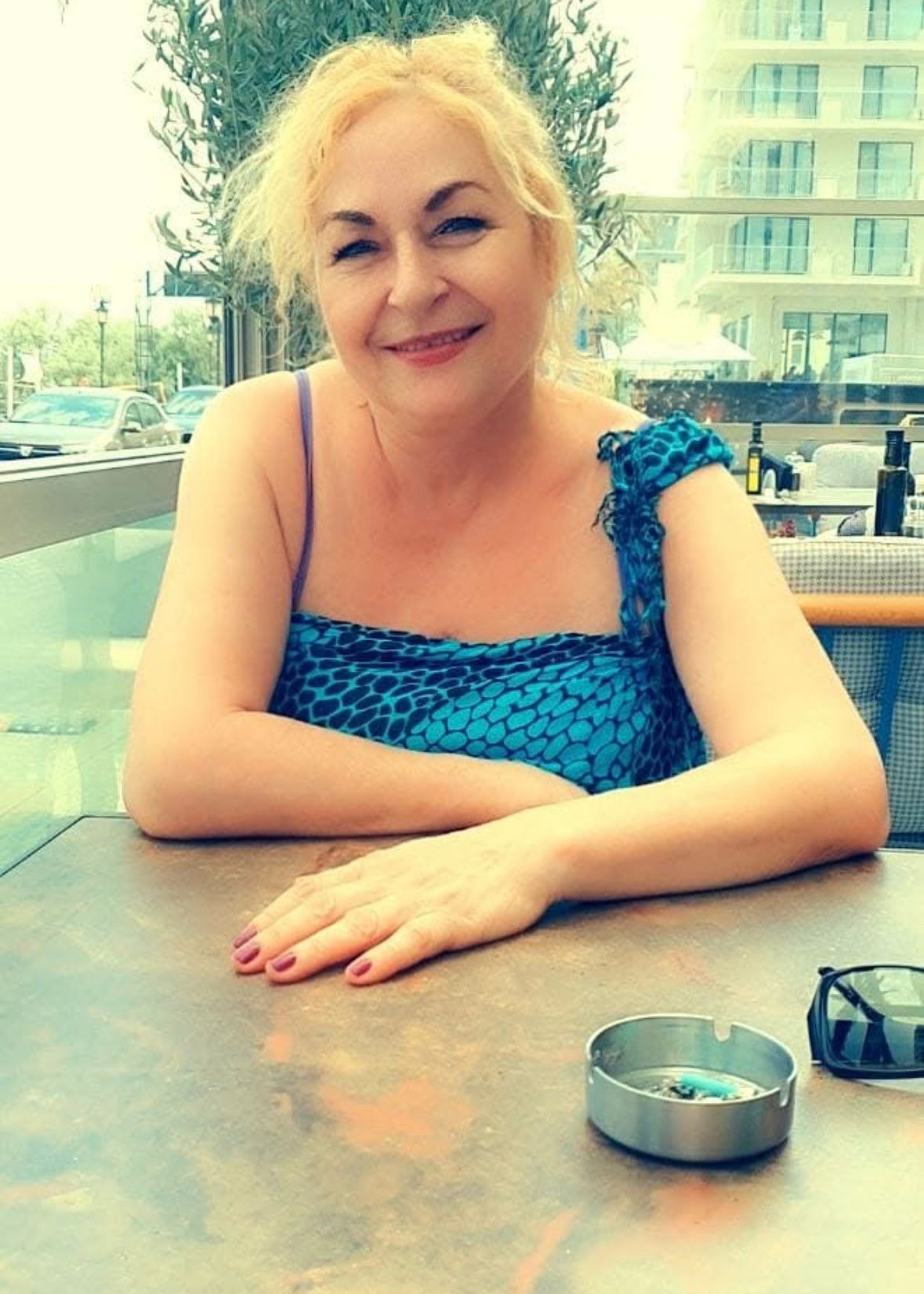 Avocata Vasilica Enache într-o rochie albastră, cu părul prins la spate, stând pe terasa unui restaurant, ținând una dintre mâini pe masă, în timp ce lângă ea se afla o scrumieră plină