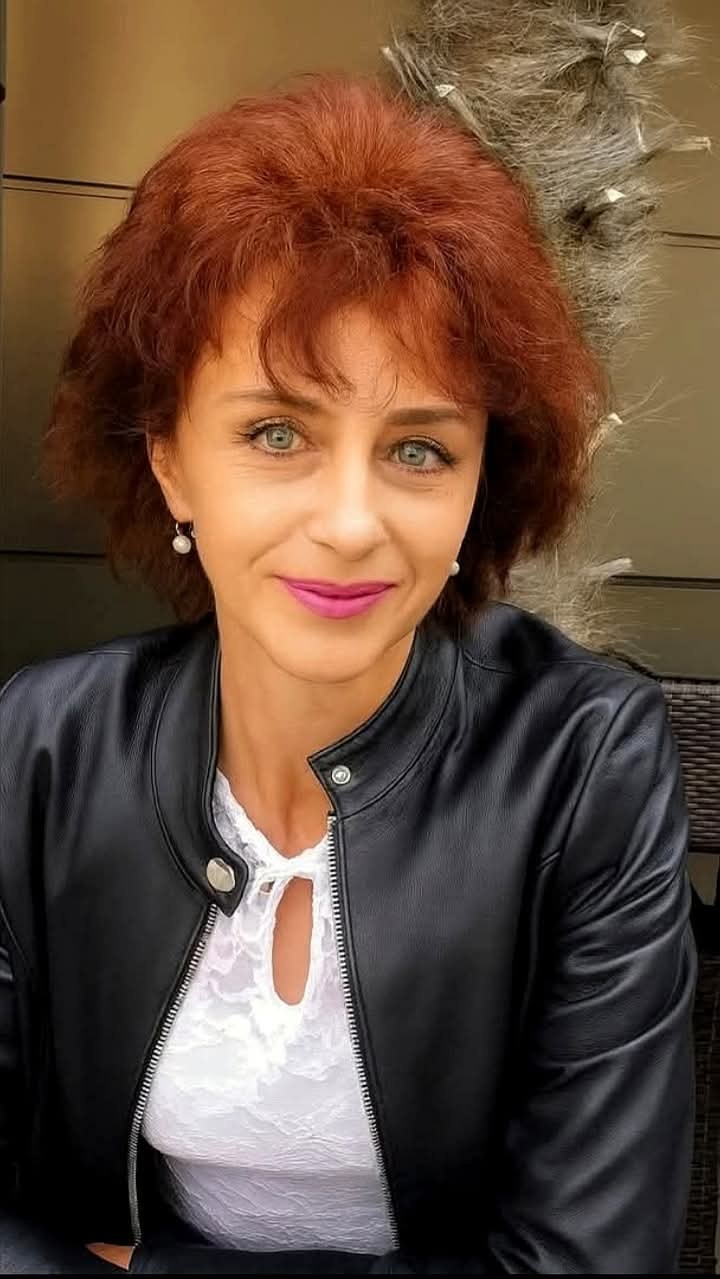 Flavia Groșan zâmbitoare, îmbrăcată în bluză albă din dantelă, cu geacă de piele și cu ruj roz pe buze