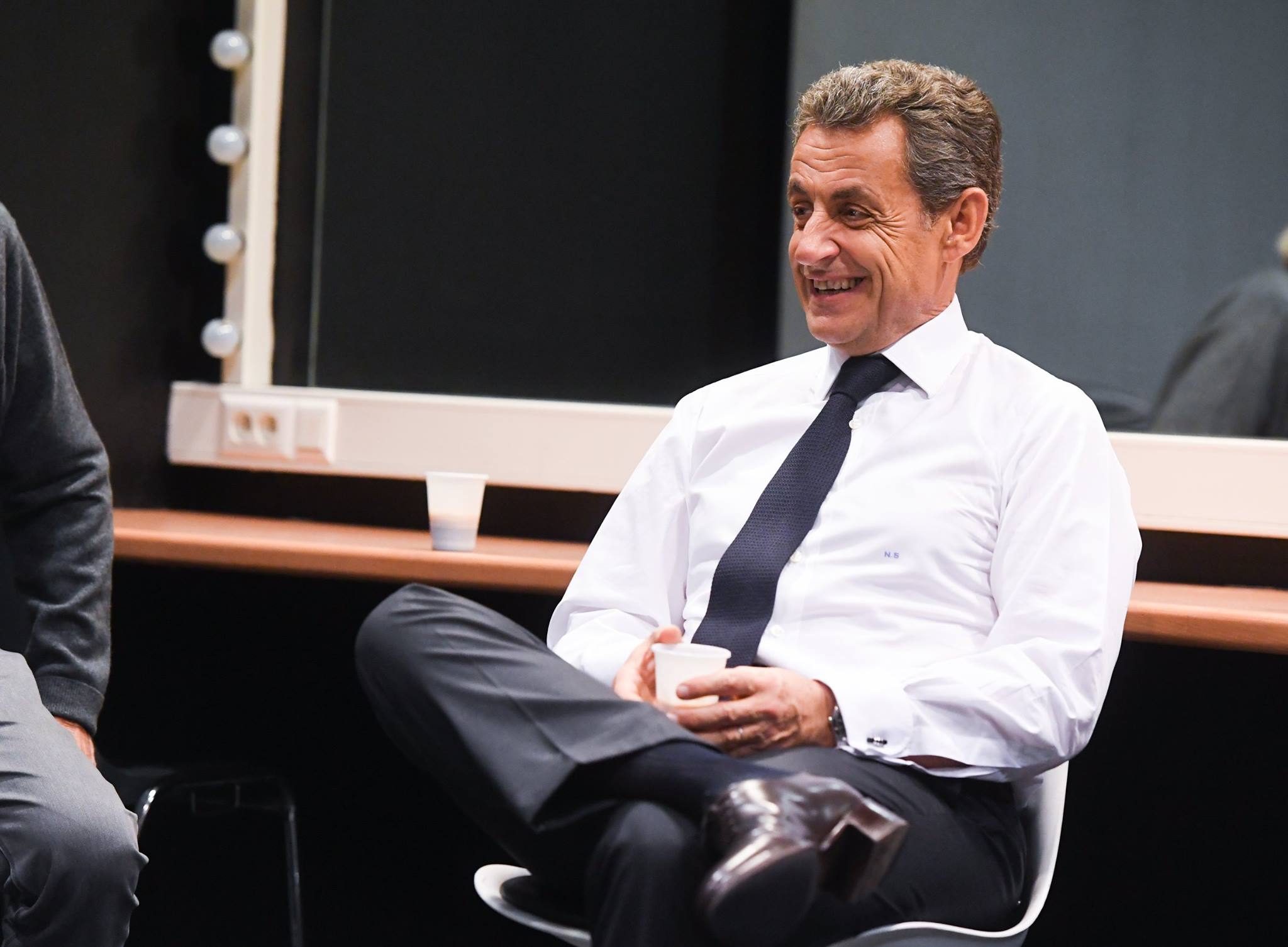 Nicolas Sarkozy, fostul președinte al Franței stând pe un scaun, zâmbitor, cu un pahar de apă în mână, îmbrăcat în pantaloni negri, cămașă albă și purtând o cravată neagră