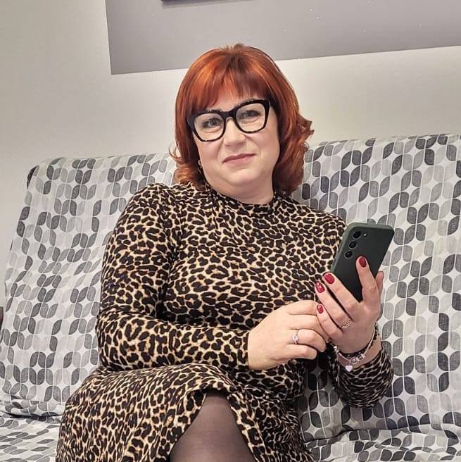 Mariana Căldăruș într-o rochie cu animal print, stând pe canapea, cu telefonul în mână și ochelarii la ochi