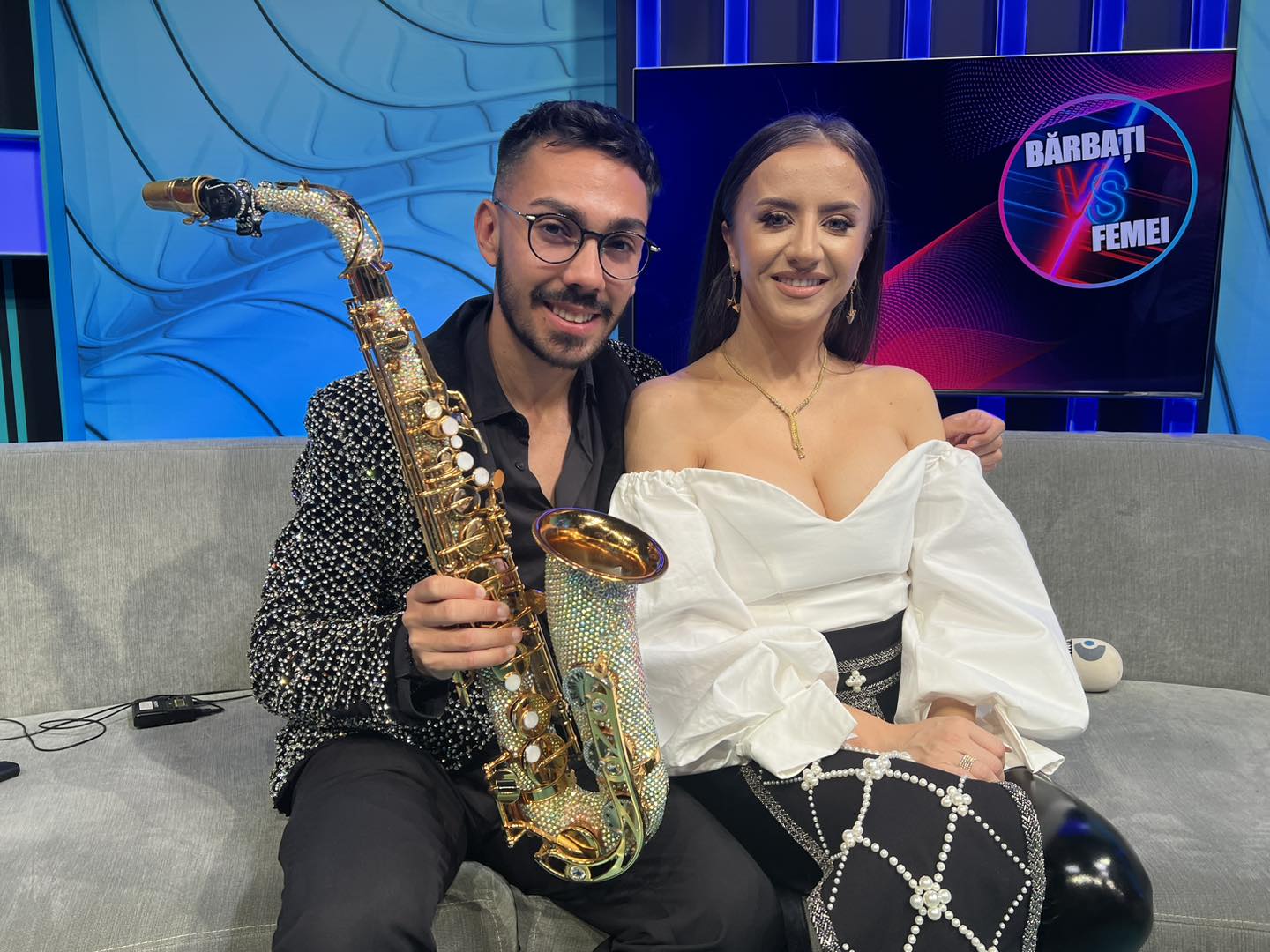 Armin Nicoară ținând saxofonul în mână, stând lângă nașa lui de cununie, Stana Stepănescu, îmbrăcată în costum popular reinterpretat, pe o canapea, în platoul unei emisiuni