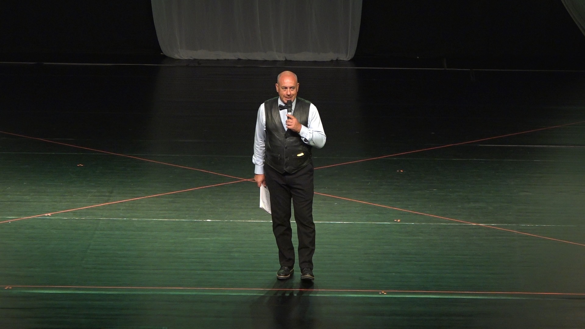 Actorul Emil Mitrache pe scenă, în timpul unui spectacol îmbrăcat în pantaloni negri, cămașă albă și vestă neagră