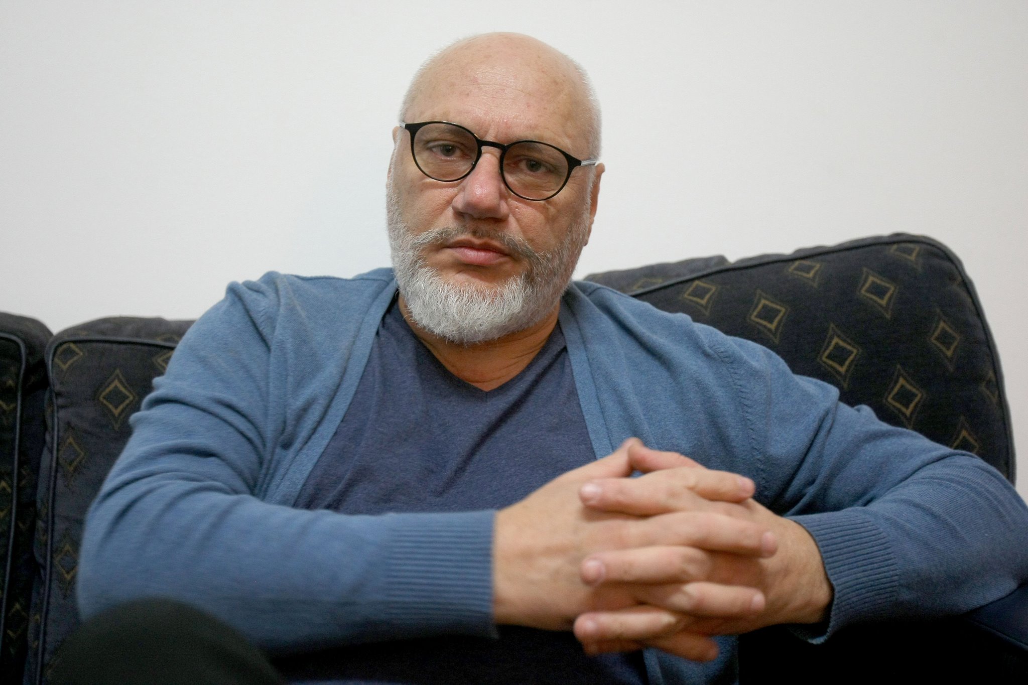 Doctorul Cristian Andrei stând serios pe o canapea, cu palmele încrucișate, îmbrăcat în nuanțe de albastru