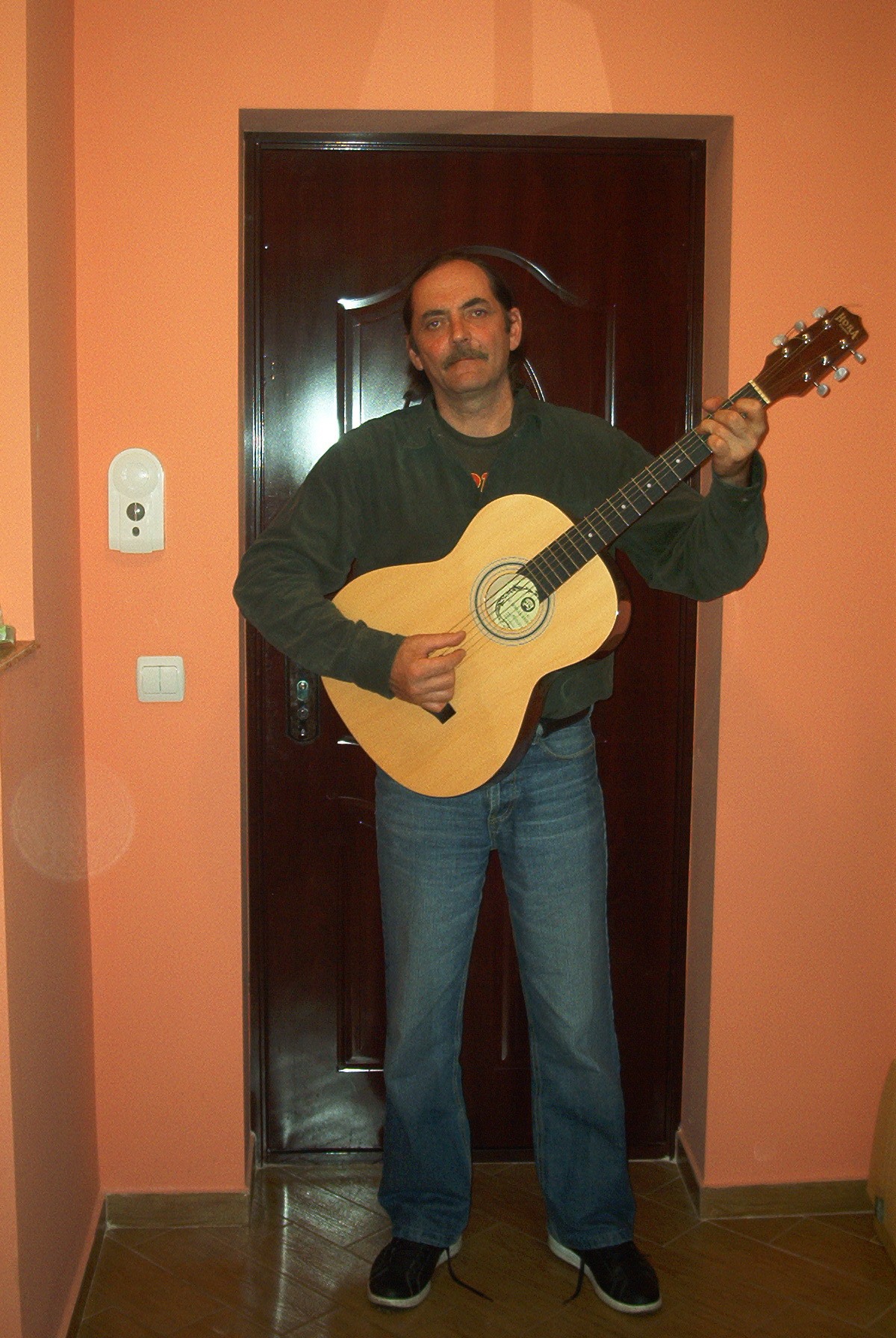 Valentin Moldovan stând lângă o ușă maro, cu chitara în mână, pregătit să cânte