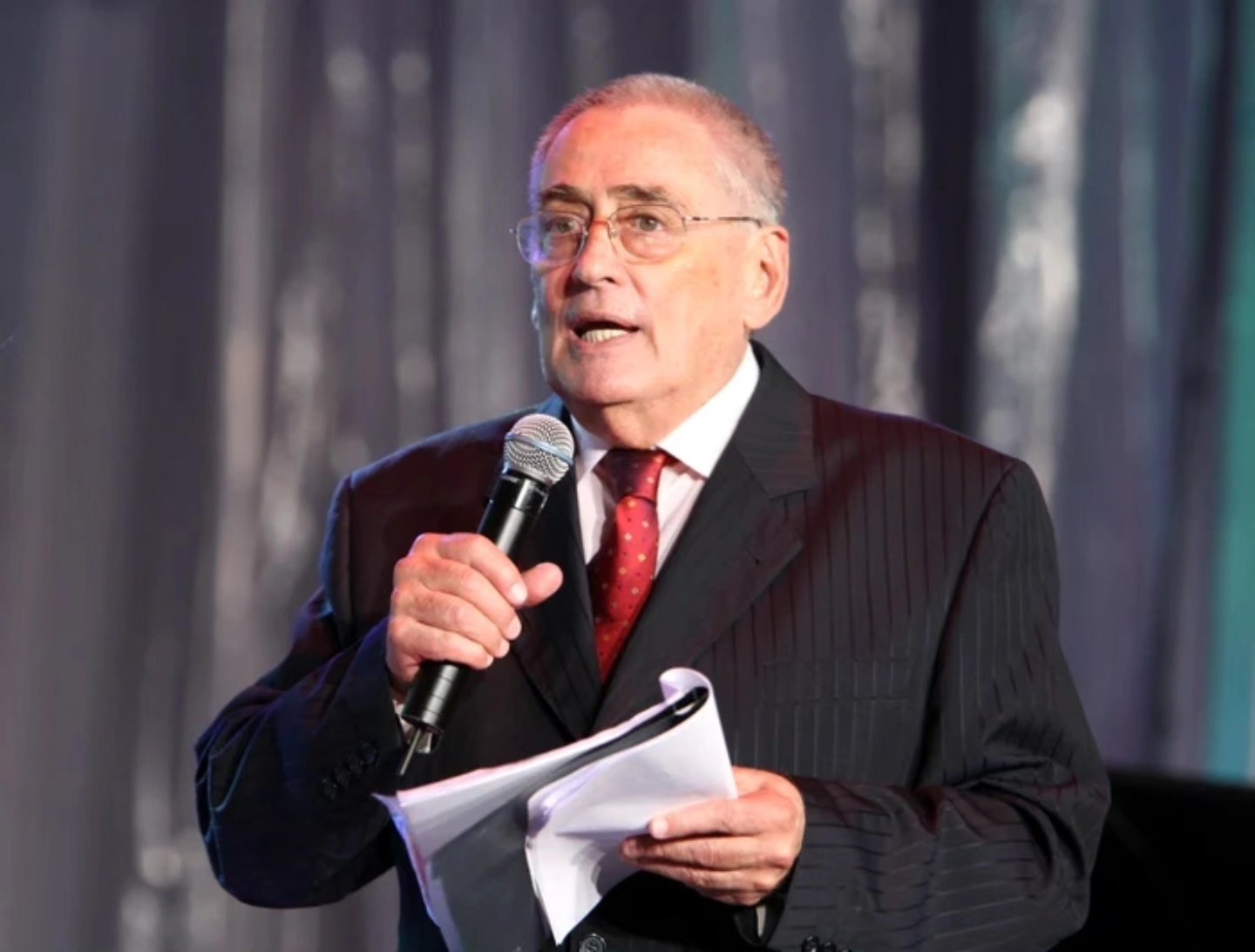 Horia Moculescu pe scenă, vorbind în fața publicului său, în costum negru, cămașă albă și cravată roșie