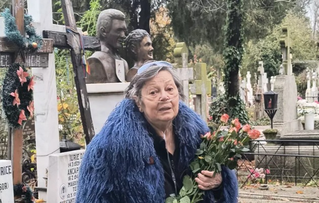 Anca Pandrea conectată la aparatul de oxigen, ținând în brațe un buchet de trandafiri pe care l-a dus la mormântul lui Iurie Darie