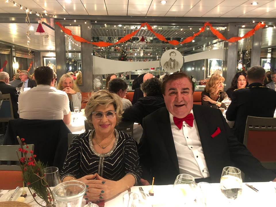 Angela Zahiu și soțul ei Ion Zahiu stând împreună la masa unui restaurant, zâmbitori, îmbrăcați elegant, ea în rochie neagră cu argintiu, el în costum negru cu cămașă albă și papion roșu