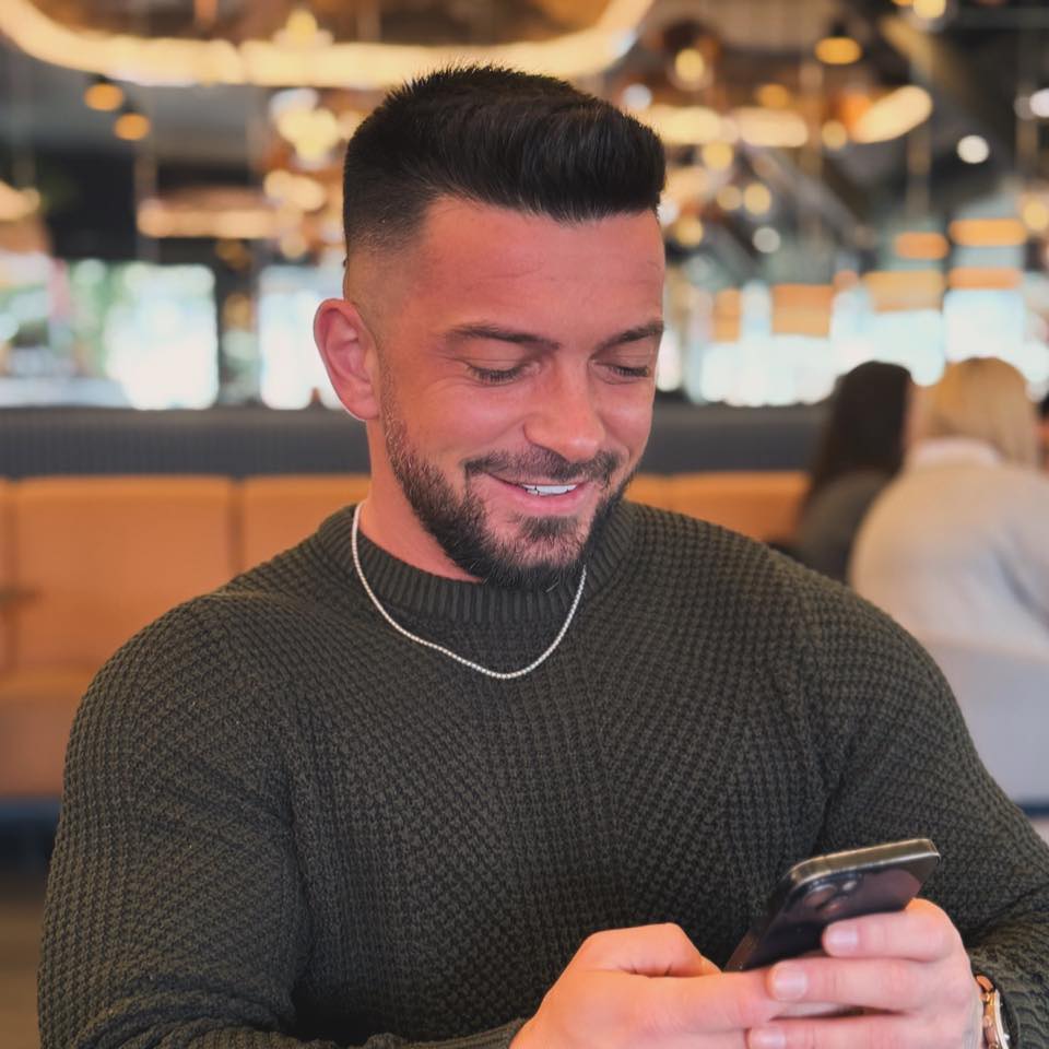 Andrei Perneș zâmbitor, uitându-se pe telefon, îmbrăcat într-un pulover kaki, cu lanț la gât și ceas la mână