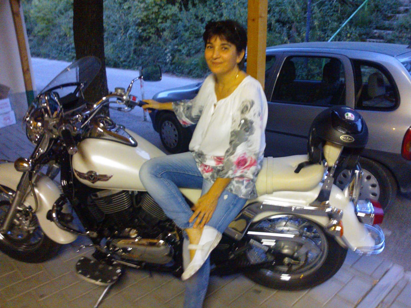 Românca Nicoleta Enache zâmbitoare, stând sprijinită de un motor, îmbrăcată în blugi deschiși la culoare și bluză albă cu flori roz